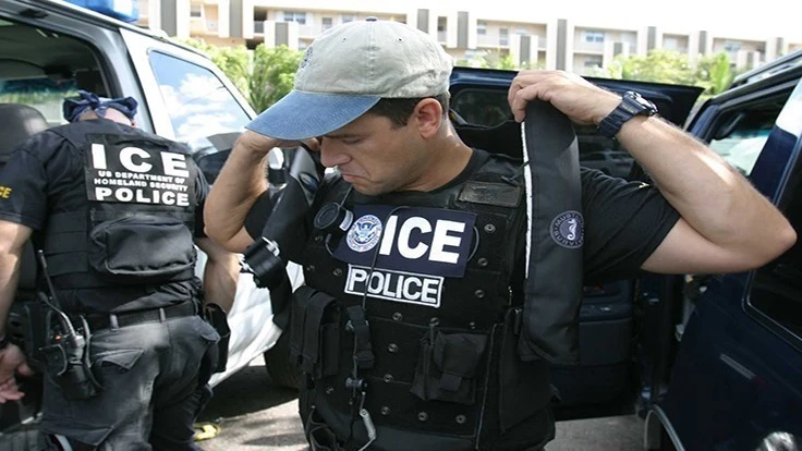 A man wearing a tan and navy blue baseball hat wearing a black shirt and black police vest that says ICE Police.